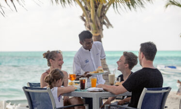 Family enjoying a beachfront meal served by a waiter under palm trees at Meeru Island Resort with turquoise waters nearby