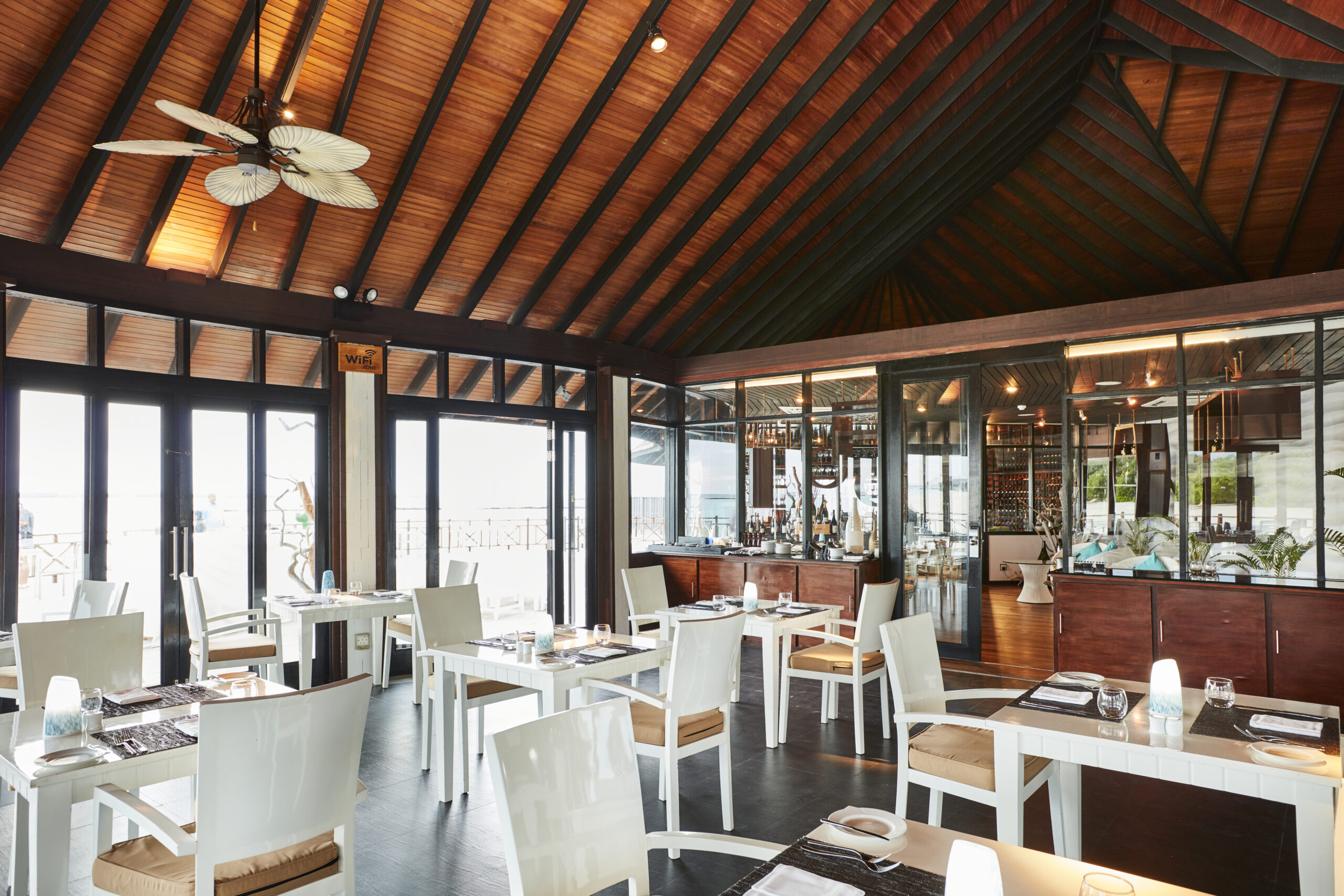 Interior of a Sun Siyam Iru Fushi restaurant featuring wooden ceilings and large glass windows with ocean views