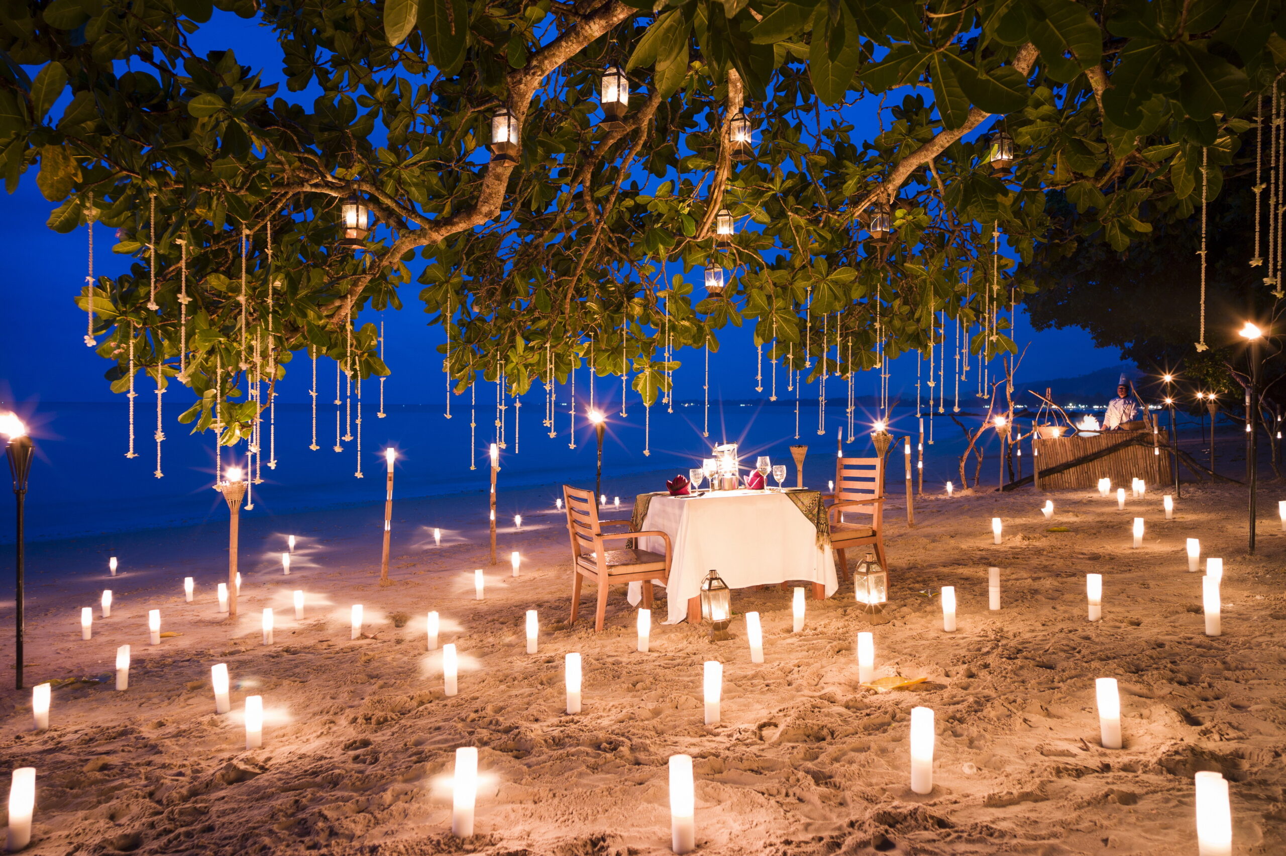 Romantic beach dinner setup with candlelit pathways under a tree at The Sarojin Khao Lak