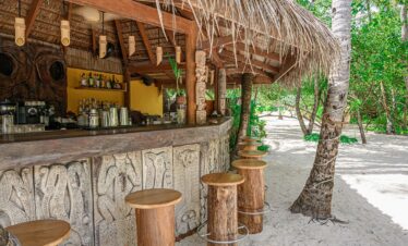 Rustic beach bar with wooden stools and tropical decor at Constance Moofushi shaded by palm trees