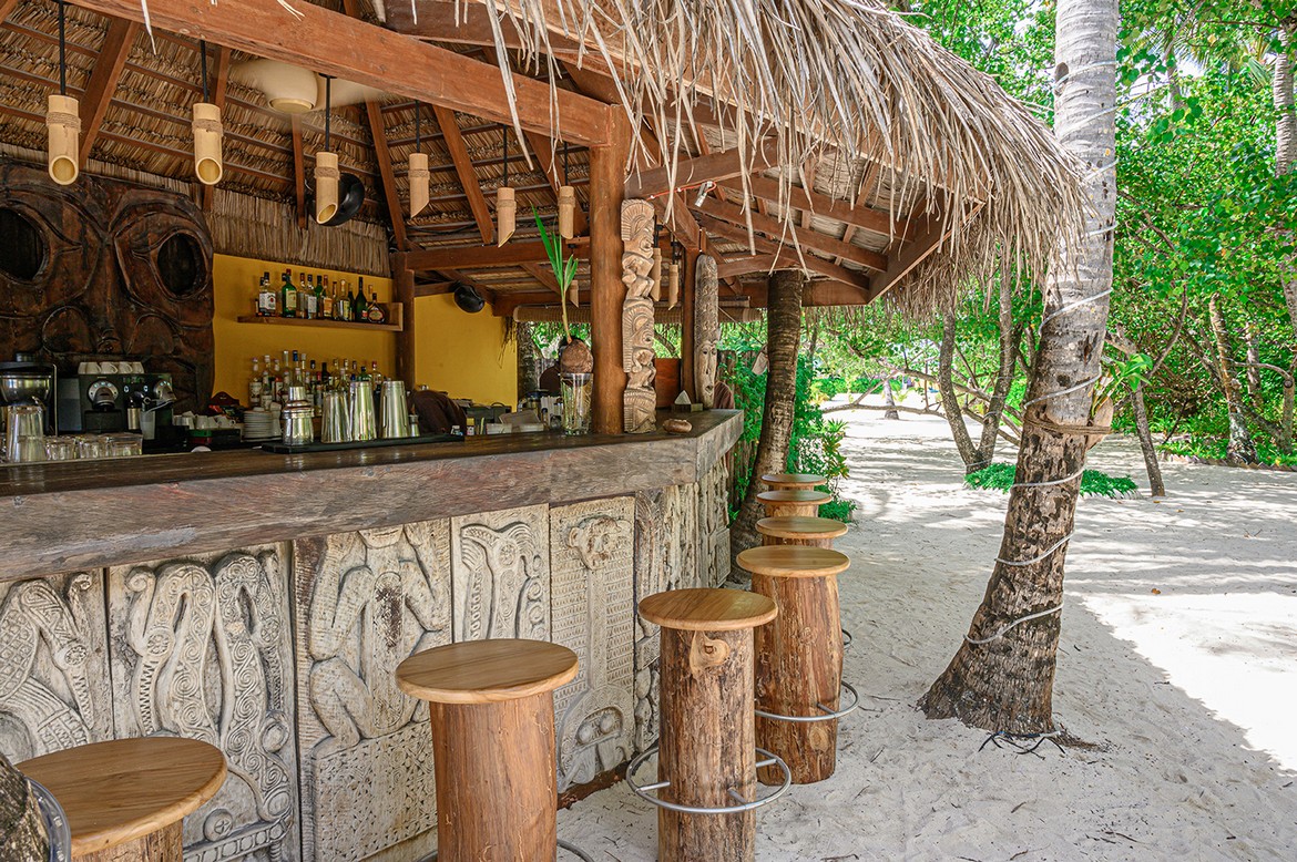 Rustic beach bar with wooden stools and tropical decor at Constance Moofushi shaded by palm trees